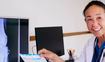 Pacientes e Cuidadores Foto de uma mulher olhando para o lado sorrindo enquanto segura uma folha de papel com informações escritas nessa folha. Ao lado dela há monitores com telas de raio-x. A mulher é adulta, asiática, de cabelo escuro preso em rabo de cavalo, veste jaleco branco com uniforme hospitalar azul por debaixo.