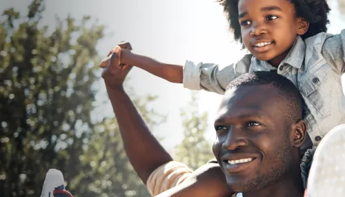 Foto de um homem sorrindo com uma criança sentada sob os seus ombros de braços erguidos para o ar, ao fundo da imagem aparece o céu e os galhos de árvores, com folhas. O homem é adulto, preto, de cabelo curto. A criança é preta, tem cabelos cacheados médio escuro, usa uma blusa social e calça jeans. Ambos olham para o horizonte num dia ensolarado. 