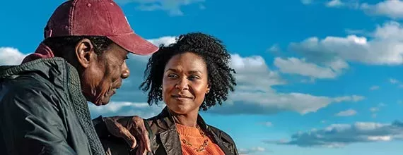 Relatório Integrado Foto de um homem e uma mulher com fundo de nuvens e céu azul. A mulher é preta com cabelo crespo médio, olha para o homem com sorriso discreto. Ela veste uma jaqueta marrom e blusa laranja. O homem, mais idoso, preto, de cabelo curto e boné vermelho, veste uma jaqueta verde cinzenta e olha para próximo da mulher.